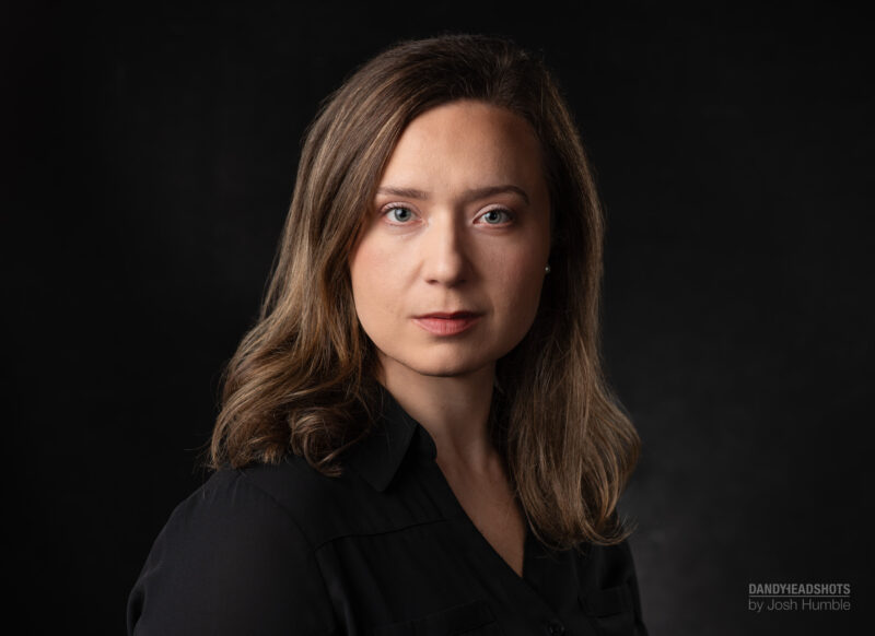 Low key headshot of woman, black backdrop, stoic facial expression.
